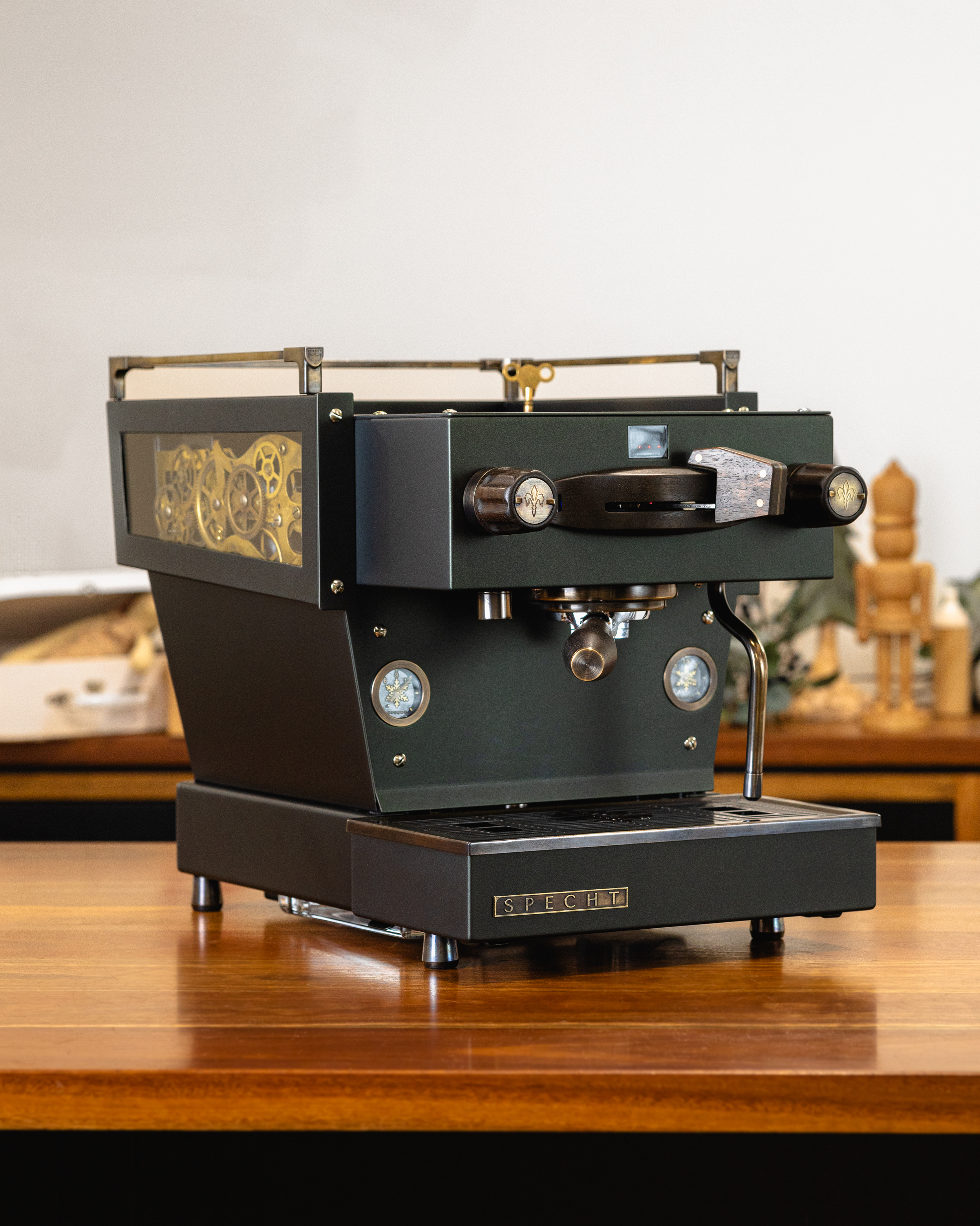 A luxurious, dark green Linea Mini R home espresso machine, reimagined by Specht with walnut touchpoints, aged brass highlights, and intricate cogs displayed through transparent sides.