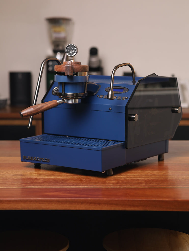A royal blue Specht La Marzocco GS3 MP with walnut wood accents and see-through, smoked glass side panels.